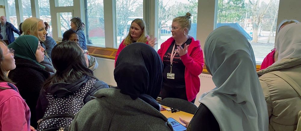 Rekryteringsmöte för vårdyrken. En ledare talar till kvinnor med huvuddukar i ett rum med stora fönster.