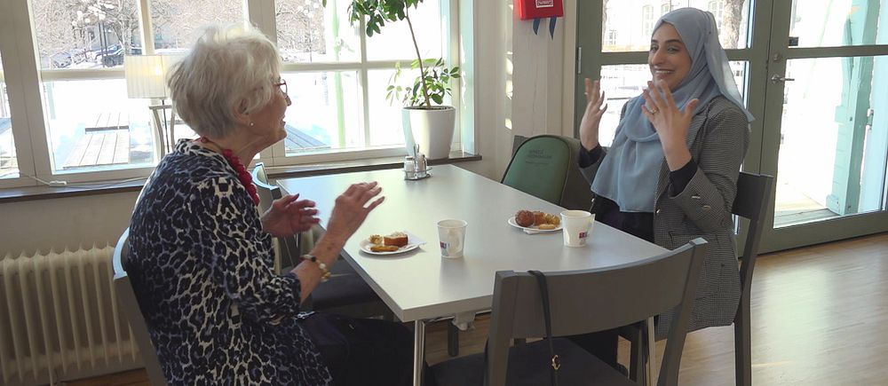 Hjördis Andersson och Sadaf Oriakhel sitter vid ett bord och samtalar över fika i ett rum med stora fönster.