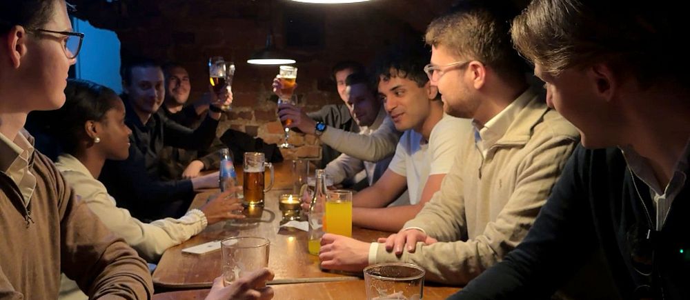 Ett större sällskap skålar och umgås runt ett långbord på en mörk pub.