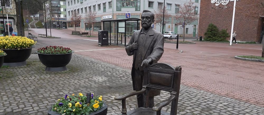 Staty av Alfred Nobel med stol på stensatt trottoar, omgiven av planteringar.
