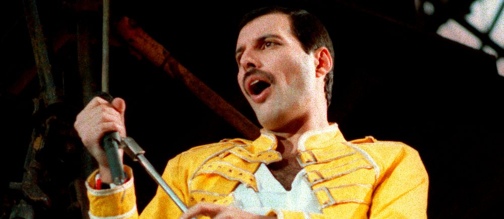 Freddie Mercury i gul jacka håller en mikrofon på scen.