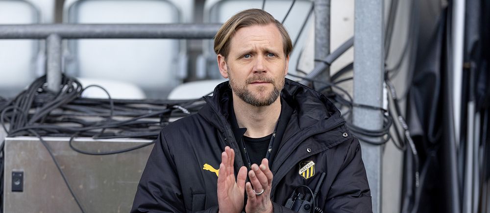 Person i svart sportjacka applåderar vid sidan av läktare med tomma stolar i bakgrunden.