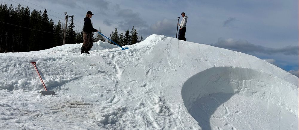 Två personer står på ett snötäckt hopp i en skidbacke, barrträd i bakgrunden.