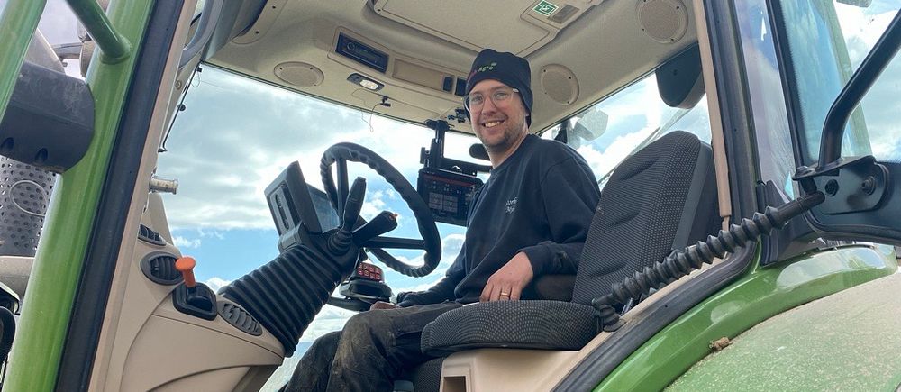 Man med mössa sitter i förarsätet på en grön traktor och ler mot kameran.
