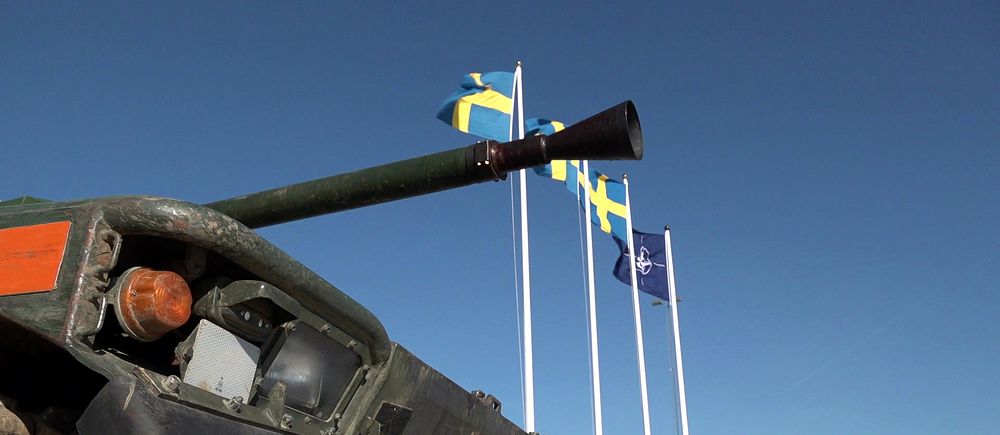 Stridsvagn framför svenska och NATO:s flaggor på klarblå himmel.