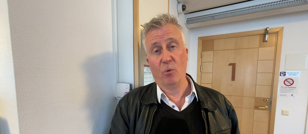 Person med grått hår håller en SVT-mikrofon i en korridor utanför en dörr märkt med en etta.