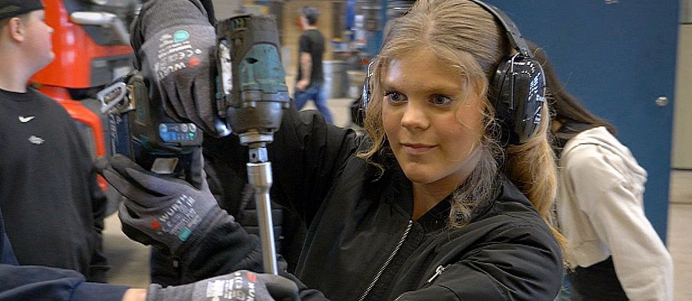 tjej arbetar med mutterdragare i verkstad, flera elever syns i bakgrunden