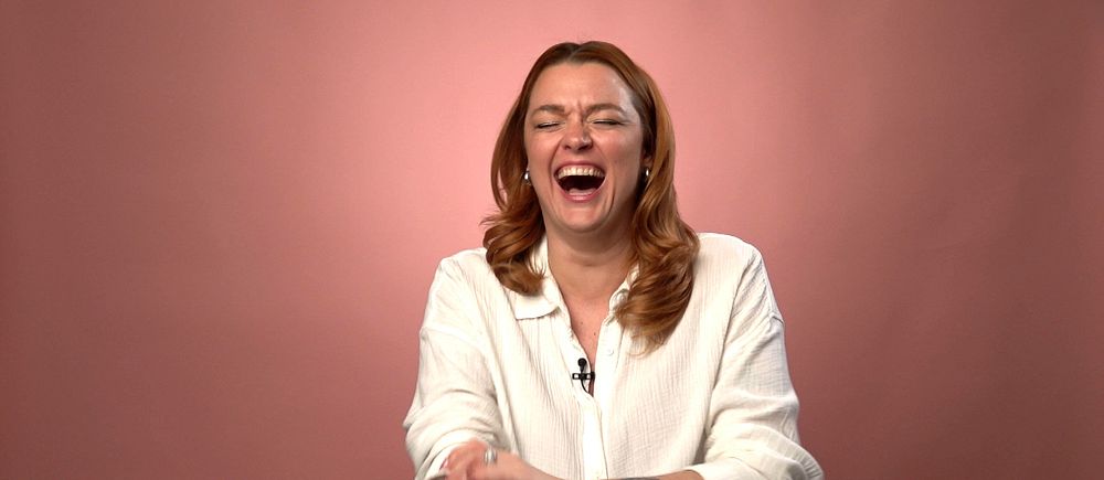 Komikern Kirsty Armstrong gapskrattar. Hon har långt rött hår och tatueringar på armena. Sitter mot en rosa bakgrund.