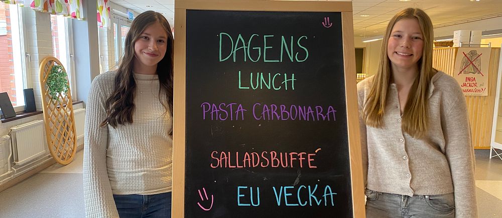 Två personer står vid en meny-tavla där det står ”Dagens lunch: Pasta carbonara, salladsbuffé, EU-vecka”.
