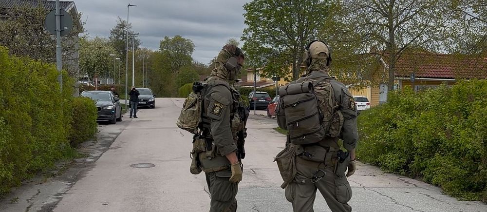 Två tungt beväpnade poliser står på en avspärrad gata i bostadsområde.