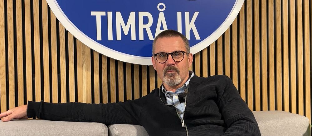 Person sitter avslappnat i soffa framför Timrå IK:s stora klubbmärke på väggen.