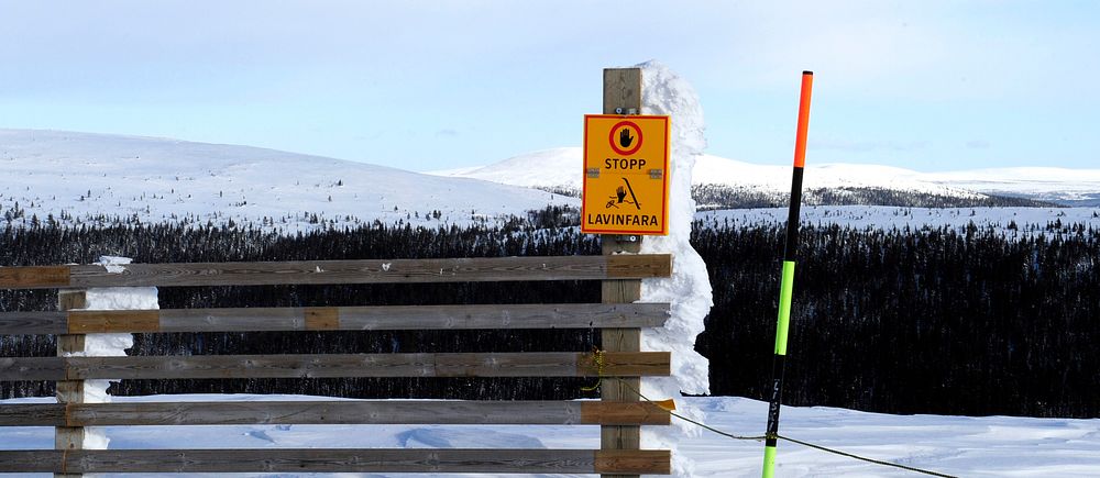 Skylt varning för lavinrisk vid snötäckt berg med trägrind och skidstolpe.