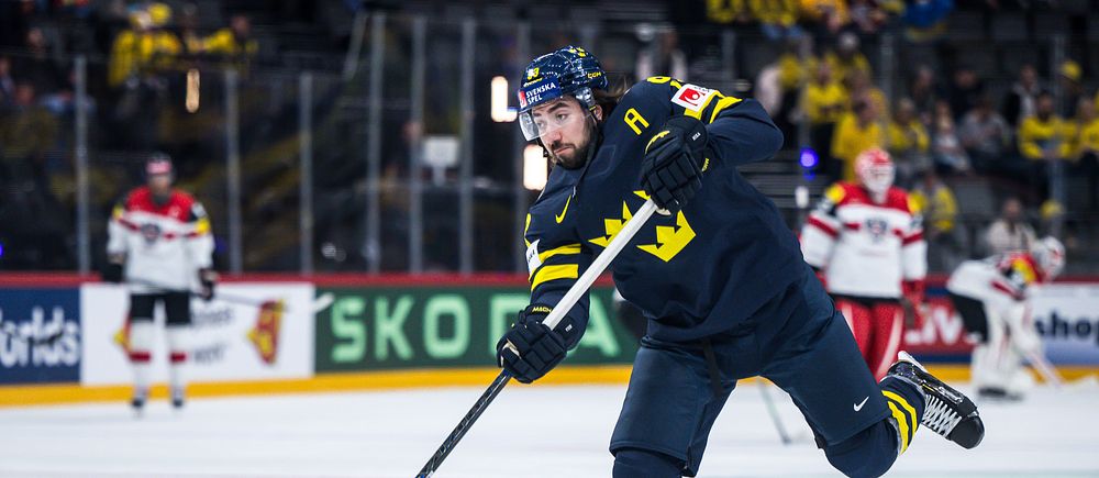 Svensk ishockeyspelare skjuter pucken under en landskamp mot lag i vita och röda tröjor.