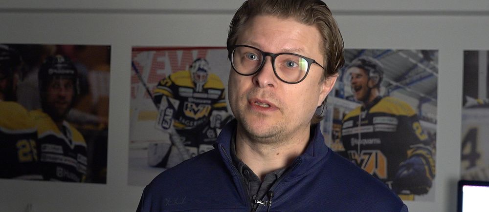 Hockeytränare med blå jacka framför tre matchfotografier på väggen.