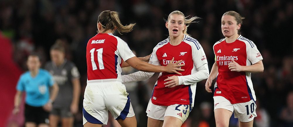 Tre kvinnliga fotbollsspelare i Arsenal-tröjor jublar på planen under en match.