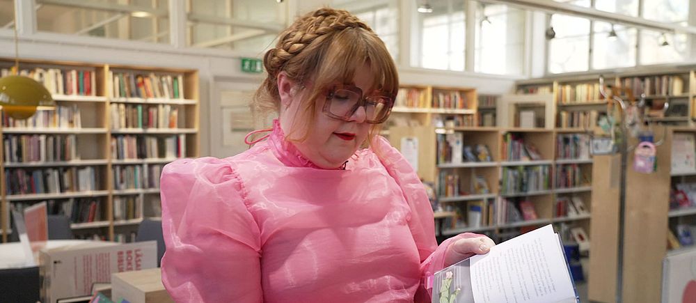 Person i rosa klänning bläddrar i bok på ett bibliotek med bokhyllor i bakgrunden.