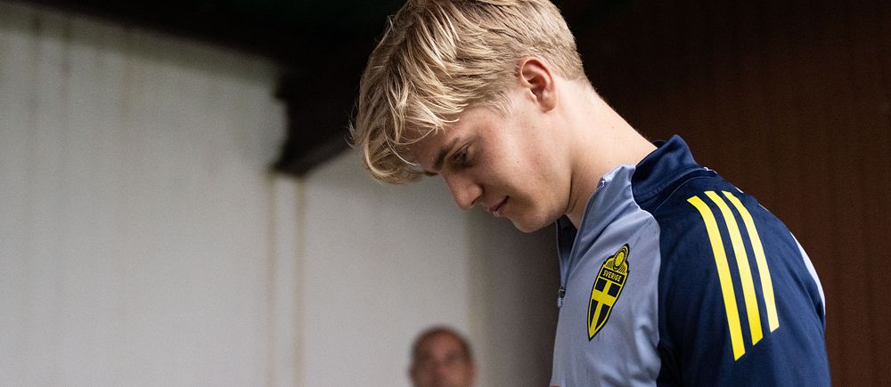 Person i svensk fotbollströja står under ett tak, betraktad av en man i bakgrunden.