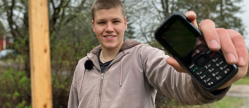 Person i beige hoodie håller fram en äldre mobiltelefon utomhus i grön miljö.