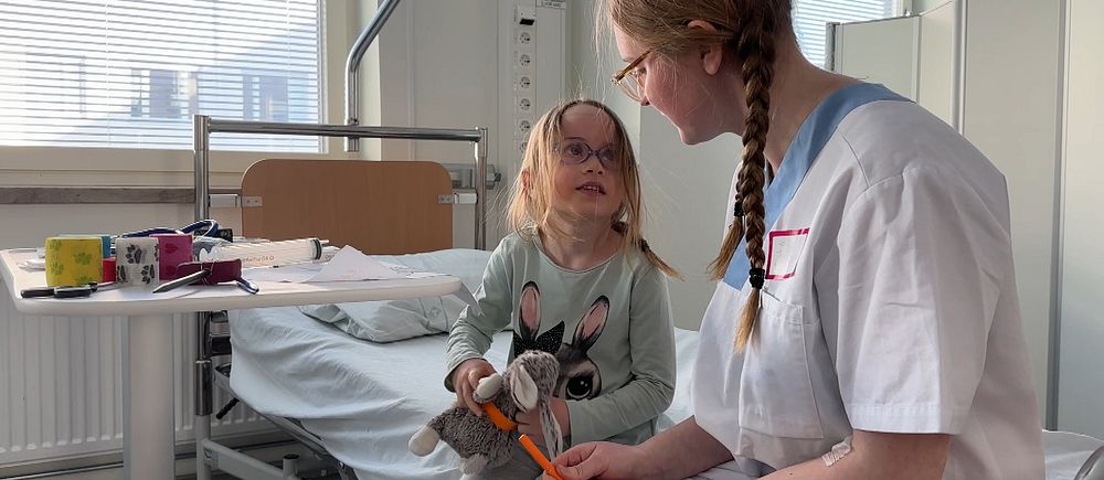 Ett barn och en vuxen kvinna sitter på en sjukhussäng och tittar på varandra. Barnet har en gosedjurskanin i handen.