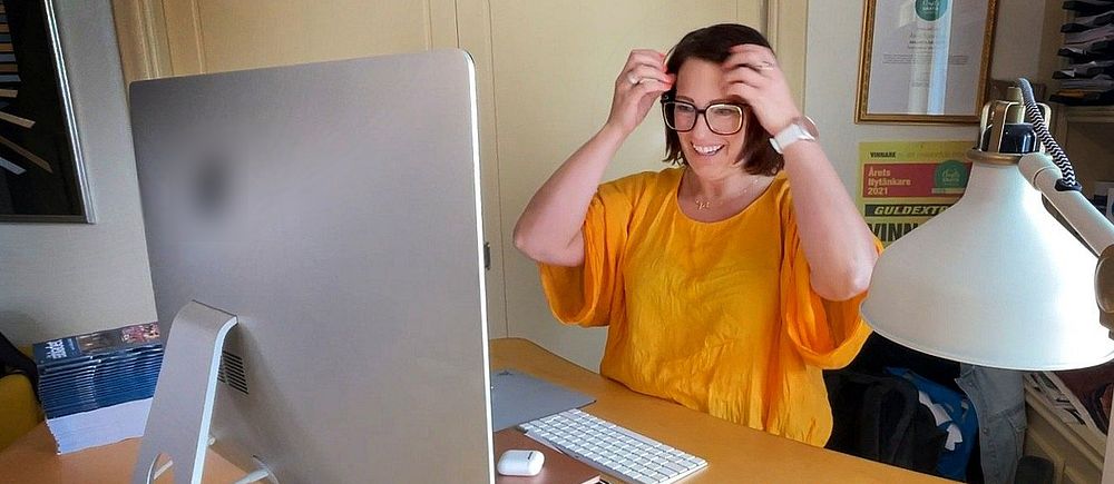 Person i gul tröja sitter vid skrivbord med dator, lyfter upp en surfplatta eller bok.