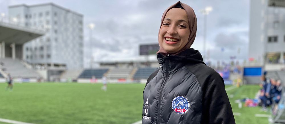 Fotbollstränare i svart jacka står vid sidan av en konstgräsplan med läktare i bakgrunden.