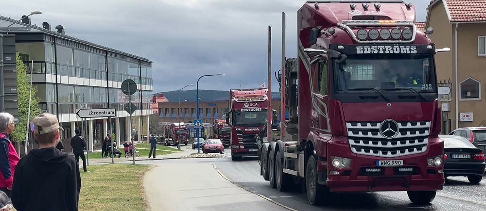 Lastbilskortege kör genom stad medan folk står vid trottoaren och tittar på.