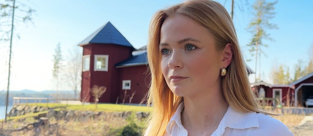 21-åriga Emma står i en utomhusmiljö under våren. Hon har på sig en vit skjorta och i bakgrunden syns ett rött hus.