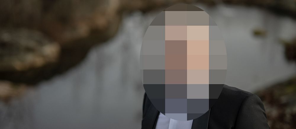 En person klädd i en mörk kostym står framför en stilla vattenspegel i en naturlig miljö.