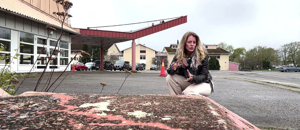 Kvinna sitter på asfalterad plats vid en övergiven byggnad och tittar mot kameran.