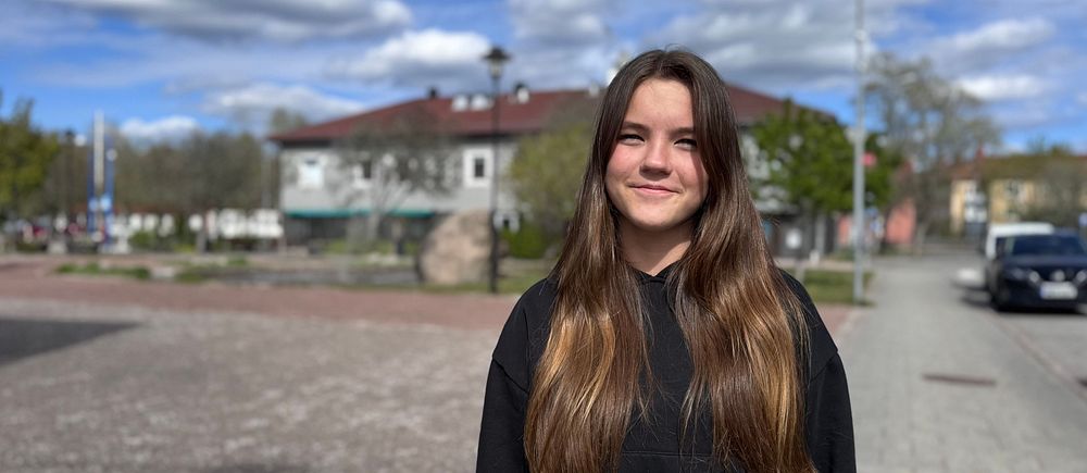 En person med långt brunt hår står på ett torg med byggnader och bilar i bakgrunden.