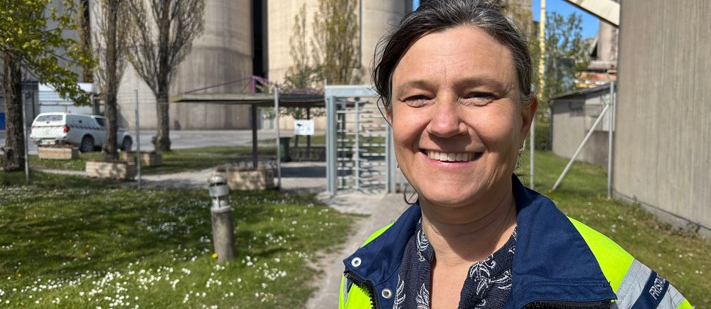 Leende person med reflexjacka utomhus vid industrimiljö med silos och grön gräsmatta.