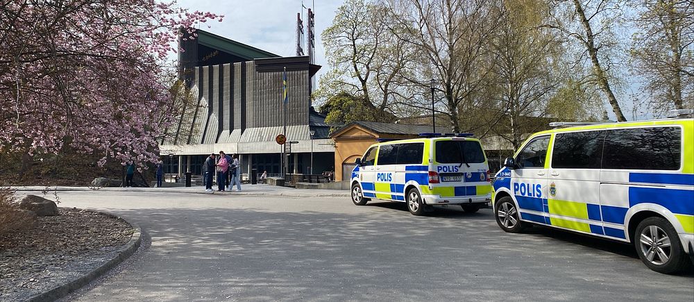 Två polisfordon vid en byggnad omgiven av träd, med människor i bakgrunden.