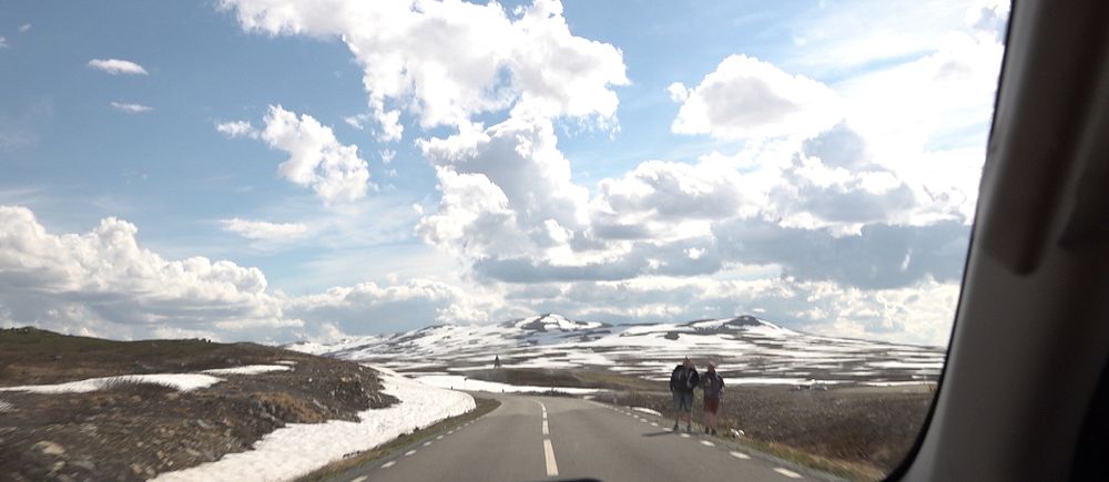 Landsväg genom fjällandskap med snö, två vandrare ses längre fram på vägen.
