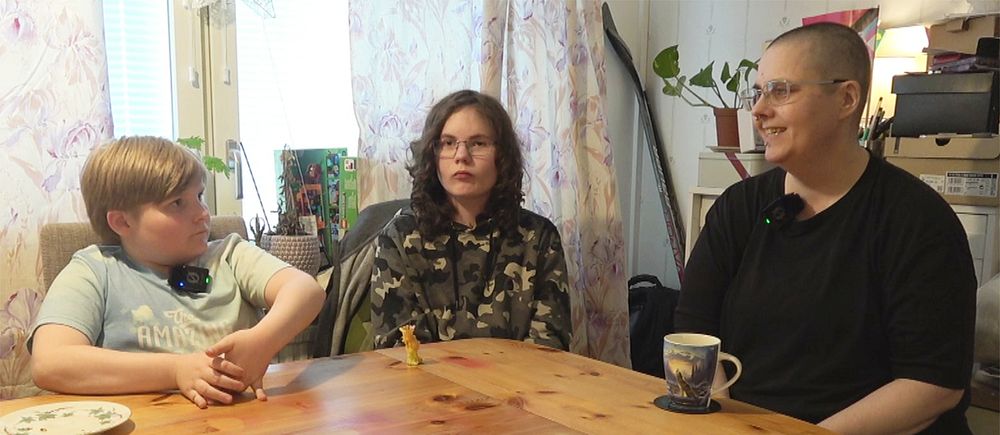 Tre personer, två pojkar och en förälder, sitter vid ett köksbord med muggar och tallrik i hemmiljö.