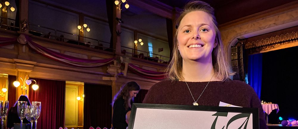 Person håller diplom i församlingshus, scen i bakgrunden.