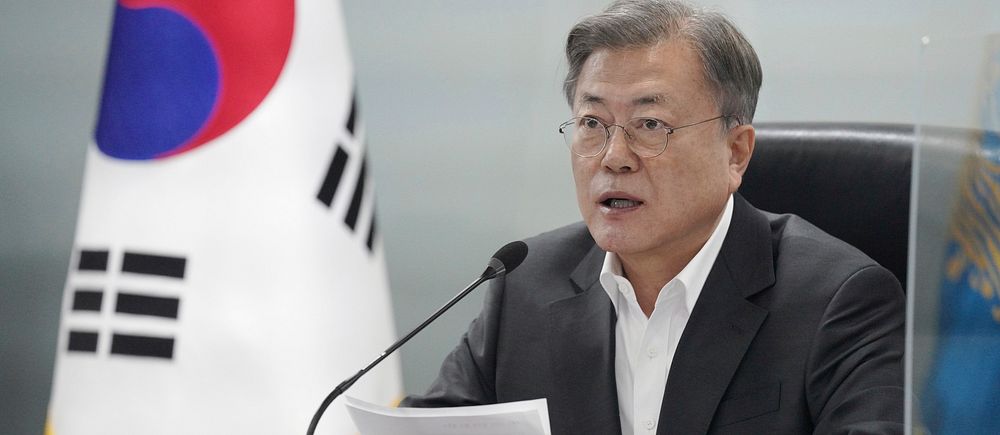 En man i grå kostym talar i en mikrofon med en sydkoreansk flagga till vänster.
