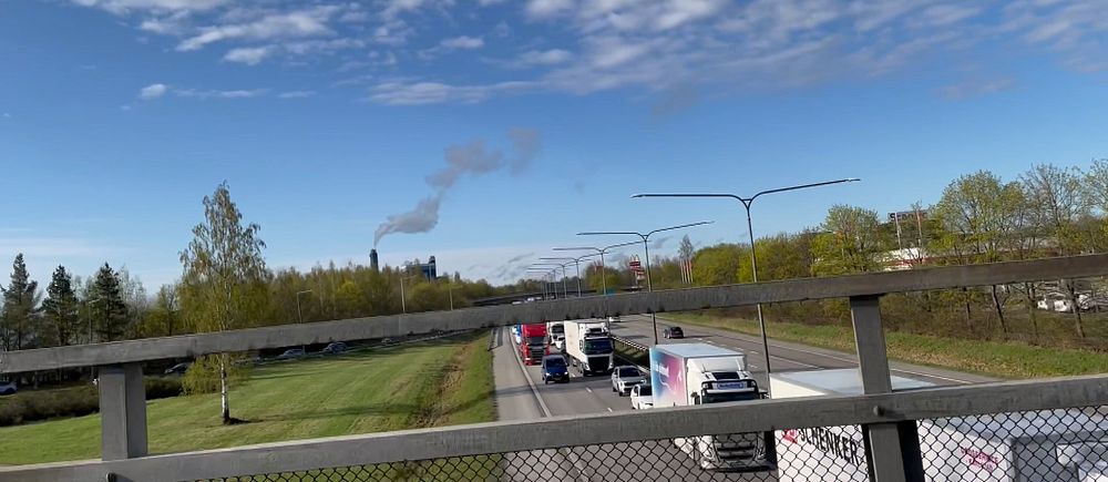 Bilkö på motorväg under blå himmel, sedd från gångbro med gröna träd på båda sidor.