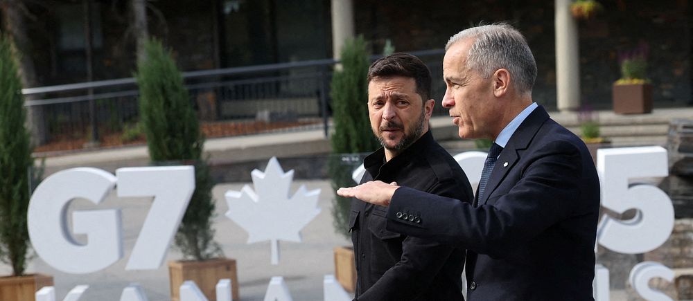 Två personer promenerar framför G7 Kanada 2018-skylt utomhus.