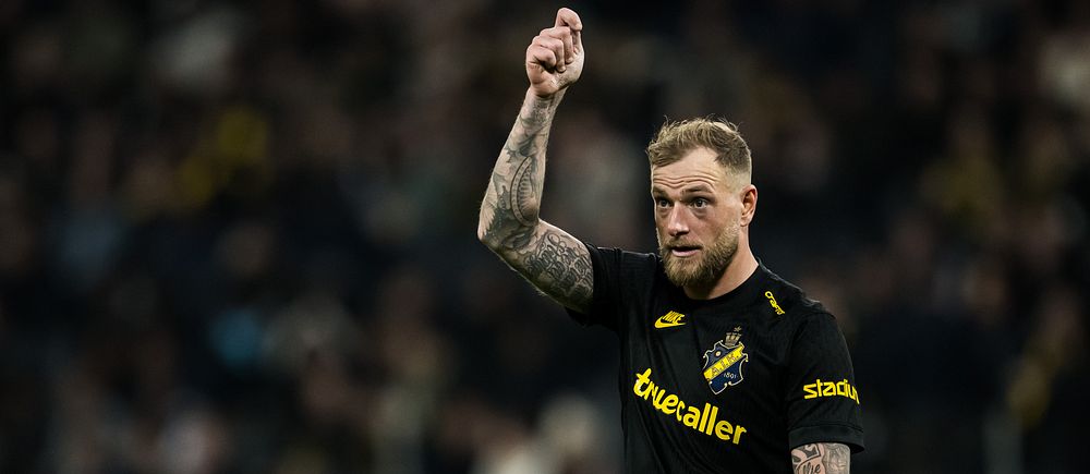 Fotbollsspelare i AIK-tröja höjer handen under match, suddiga åskådare i bakgrunden.