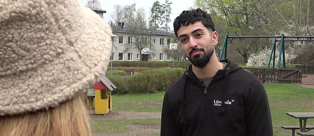 En man i svarta kläder står på en förskolegård. En kvinna med hatt står i förgrunden.