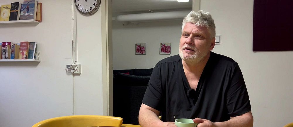 Man med grått hår sitter vid ett bord med en mugg i en vardagsrumsmiljö.