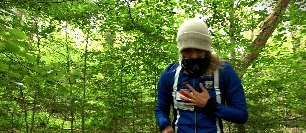 Sara Hagström med mössa, buff och tre jackor i skogen mitt i sommaren.