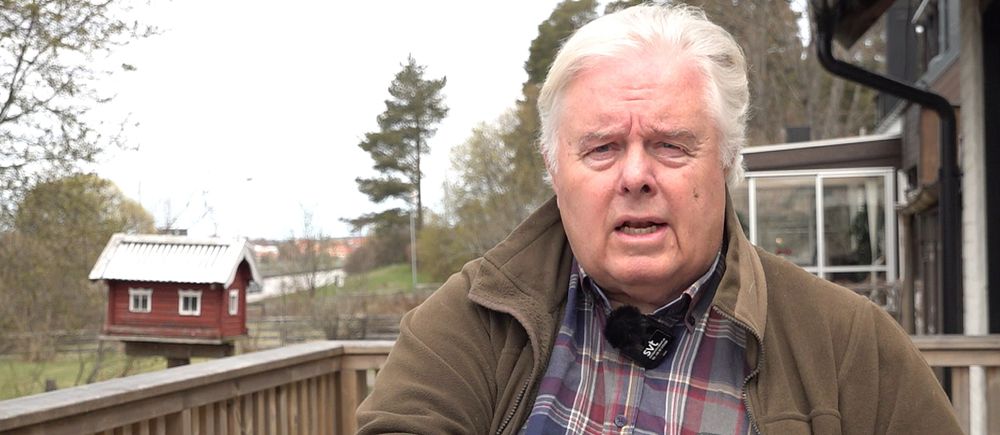 En person i rutskjorta sitter utomhus vid ett trästaket, med en röd fågelholk i bakgrunden.