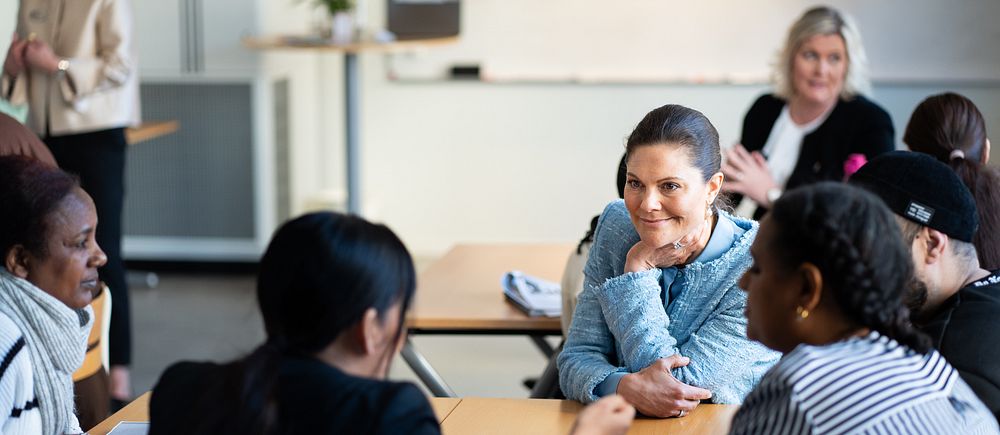 Elever sitter vid ett bord med kronprinsessan Victoria och diskuterar i ett klassrum, med landshövding Lena Rådström Baastad i bakgrunden.