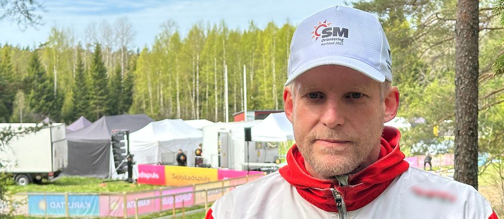 Person i vit GMOK-mössa och jacka vid ett utomhusevenemang i skogsmiljö.
