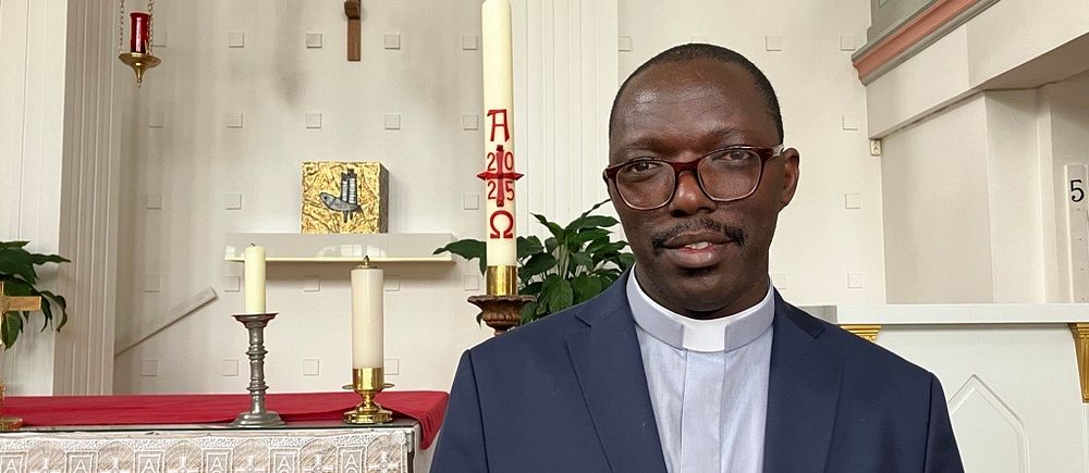 Präst i kavaj står framför altaret i en kyrka med kors och ljus i bakgrunden.