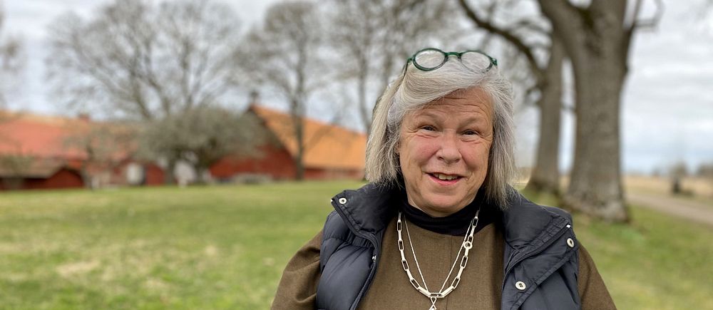 Kvinna med grått hår iklädd tröja och täckväst står utomhus, tittar in i kameran. I bakgrunden syns träd och en ladugård.