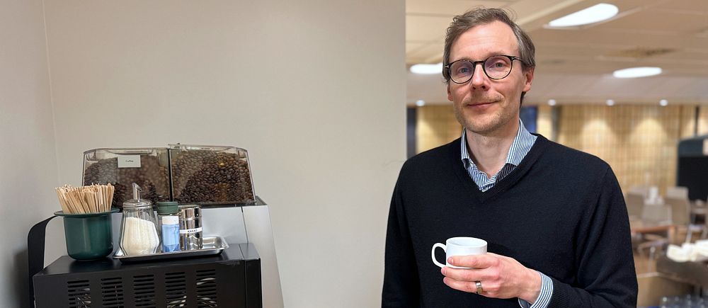 Person står bredvid en kaffemaskin, håller en liten kaffekopp. Bakgrund med bord och stolar.