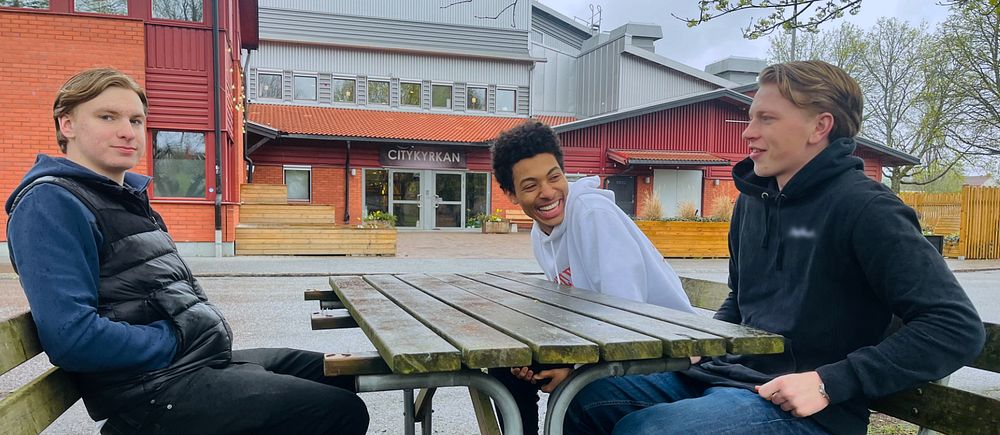 Tre personer sitter vid ett utomhusbord framför en byggnad. En av dem skrattar.