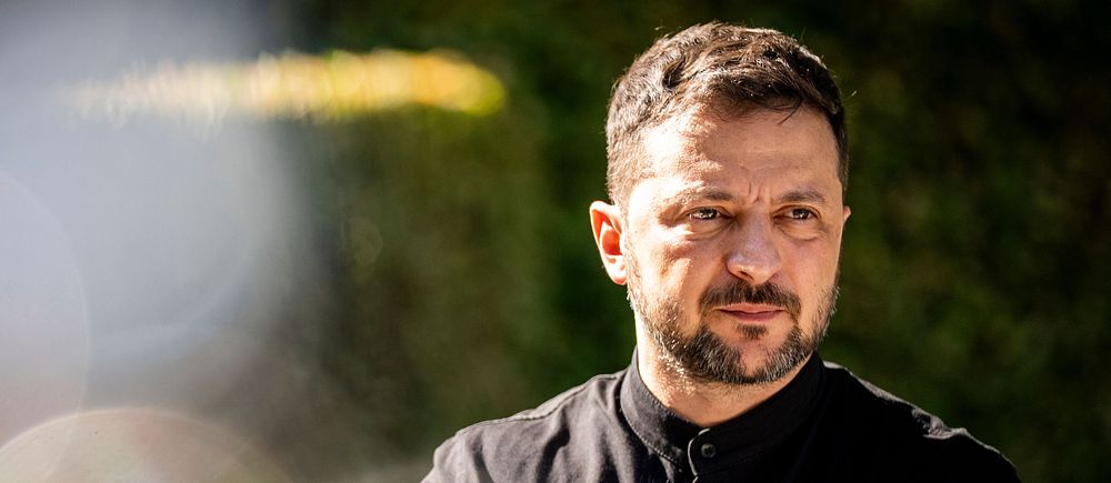 Zelenskyj i svart tröja utomhus med suddig bakgrund och ljusreflexer i förgrunden.